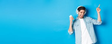Mutlu yakışıklı adam dans ediyor, kulaklıkla akıllı telefondan müzik dinliyor, mavi arka planda duruyor..