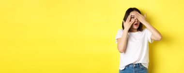 Şaşırmış Asyalı kız şaşırmayı bekliyor, parmaklarının arasından bakıyor, mutlu bir şekilde gülümsüyor, sarı arka planda duruyor..