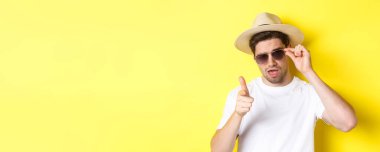 Turizm ve tatil kavramı. Havalı ve küstah bir adam seninle flört ediyor, güneş gözlüğü takıyor, kamerayı işaret ediyor, sarı arka planda duruyor..