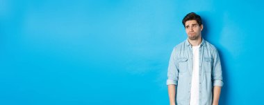 Gayri resmi giyinmiş, üzgün ve isteksiz bir adam sol tarafa bakıyor, somurtuyor ve üzgün hissediyor, mavi arka planda duruyor..