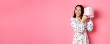 Şirin Asyalı kadın hediye kutusunu sallıyor, gülümsüyor ve ilgi çekiyor, tahmin et bakalım içeride ne var, pembe arka planda duruyor..