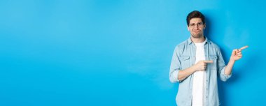 Gözlüklü genç adam tam fotokopi alanını işaret ediyor, reklam afişini gösteriyor ve tatminsiz sırıtıyor, kötü ürünü yargılıyor, mavi arka planda duruyor..