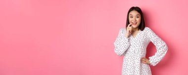 Elbiseli, ısıran, gülümseyen, baştan çıkarıcı bakışlar atan, pembe arka planda duran, bir şeyler almak isteyen güzel bir genç kadın görüntüsü..