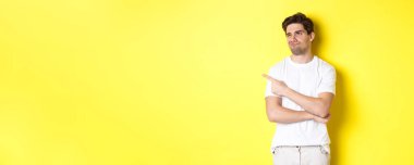 Etkilenmemiş genç adam şikayet ediyor, şüpheli bir ifadeyle kötü ürüne bakıyor, sarı arka planda duruyor..