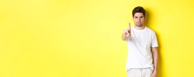 Hayır diyen kendine güvenen bir adam, bir şeyi reddeder ve yasaklar, bir parmağını gösterir ve kaşlarını çatar, sarı arka planda durur..