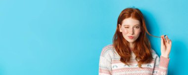 Küstah kızıl saçlı, saçlarıyla oynayan, kameraya göz kırpan ve gülümseyen, mavi arka planda duran kızın yakın çekimi..