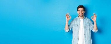 Kendine güvenen yetişkin bir adam, tamam işaretleri gösteriyor ve memnun görünüyor, bir şey gibi, mavi arka planda duruyor..