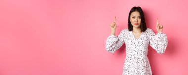 Üzgün Asyalı bir kız işaret ediyor, reklam afişini gösteriyor, hayal kırıklığına uğramış kasvetli bir bakışla kameraya bakıyor, pembe arka planda giyinmiş duruyor..