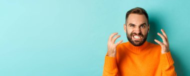 Kızgın beyaz adamın yakın plan fotoğrafı. Deli gibi bakıyor, yüzünü buruşturuyor ve el sıkışıyor. Kırılmış, açık mavi arka planda duruyor..