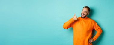 Mutlu erkek model baş parmağını gösteriyor, sol üst köşedeki logodan memnun görünüyor, ürünü onaylıyor ve tavsiye ediyor, açık mavi arka planda duruyor..