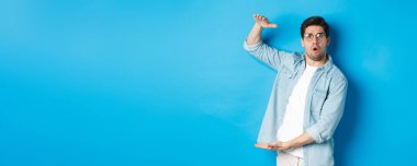 Kafası karışmış yakışıklı adam büyük boy gösteriyor, büyük kutuyu şekillendiriyor ve şaşırmış görünüyor, mavi arka planda duruyor..