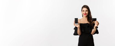 İnternetten alışveriş konsepti. Mobil ekran ve kredi kartı gösteren siyah elbiseli şık esmer kadın, gülümsüyor, beyaz arka planda duruyor..