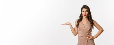 Zarif elbiseli rahatsız edilmemiş genç bir kadın neyin yanlış olduğunu anlamıyor, elini kaldırıyor ve dikkatsiz görünüyor, beyaz arka planda duruyor..