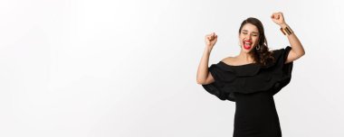 Moda ve güzellik. Siyah elbiseli başarılı güzel kadın, kazanmanın sevinci, beyaz arka planda zafer..