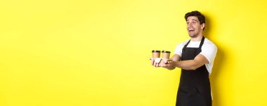 Siyah önlüklü arkadaş canlısı barista iki fincan kahve tutuyor ve gülümsüyor, sarı arka planda duruyor..