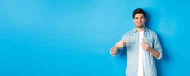 Çevrimiçi alışveriş, uygulama ve teknoloji kavramı. Şüpheci ve hoşnutsuz adam akıllı telefonu işaret ediyor ve hayal kırıklığına uğruyor, mavi arka planda dikiliyor..