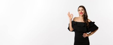 Moda ve güzellik. Kırmızı rujlu, siyah elbiseli, tatmin olmuş güzel bir kadın. Onaylama işareti var. Beyaz arka planda duruyor..