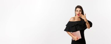 Moda ve alışveriş konsepti. Cazip makyajlı, siyah elbiseli, elinde dijital tablet olan ve şaşırmış görünen, beyaz arka planı olan şık genç bir kadın..