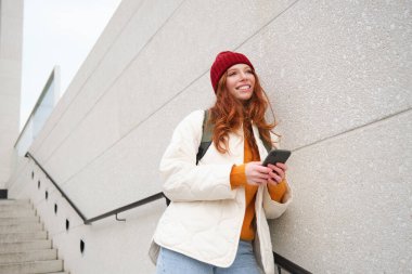 Happy girl student in red hat, holds smartphone, tourist looks at map app on her phone, explores sightseeing, texts message, looks for couchsurfing, rents place to stay online.