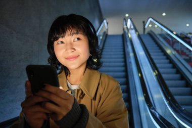 Asyalı esmer kızın gülümseyen portresi, yürüyen merdivenden inen, akıllı telefon tutan ve kulaklıkla müzik dinleyen, toplu taşıma araçlarını kullanan, şehirde seyahat eden.