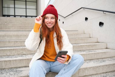 Güzel kızıl saçlı kız, öğrenci cep telefonuyla kampüsün yanındaki merdivenlerde oturuyor, gülümsüyor, kırmızı şapka takıyor, akıllı telefon uygulaması kullanıyor..
