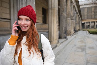 Smiling redhead female tourist talks on mobile phone and walks around city. Happy student in red hat calls friend, stands on street and uses smartphone.