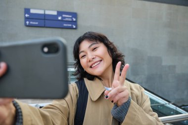 Portrait of stylish korean girl, student takes selfie on street, holds smartphone and poses on its camera, makes video on tourist trip, smiles with joy.