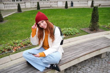 Young smiling redhead girl sits on bench and uses smartphone app, reads news online, watches video on mobile phone while relaxed in park.