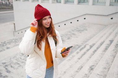Joyful girl tourist looks at mobile phone, texts message on smartphone social media application, walks around city, looks for sightseeing on mobile app.