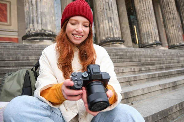 Portrait of young photographer girl, sits on stairs with professional camera, takes photos outdoors, making lifestyle shooting.