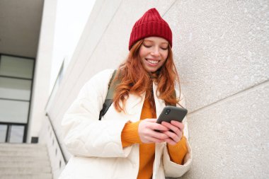 Happy girl student in red hat, holds smartphone, tourist looks at map app on her phone, explores sightseeing, texts message, looks for couchsurfing, rents place to stay online.