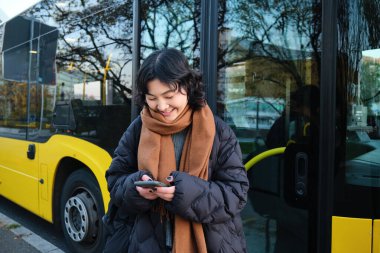 Beautiful korean girl, student on bus stop, looking at her smartphone, checking timetable, reading text message, wearing winter clothes.