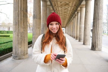 Mobile broadband and people. Smiling redhead 20s girl with backpack, uses smartphone on street, holds mobile phone and looks at application.
