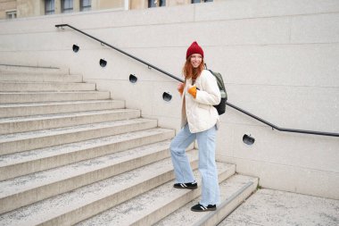 Young stylish girl traveller goes sightseeing, travels around city with backpack, tourist smiles and looks around. Copy space