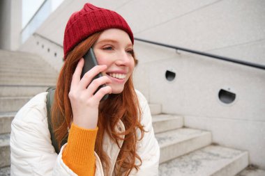 Young stylish redhead girl in red hat, sits on street and talks on mobile phone, has telephone conversation, rings her friend while relaxes outdoors.