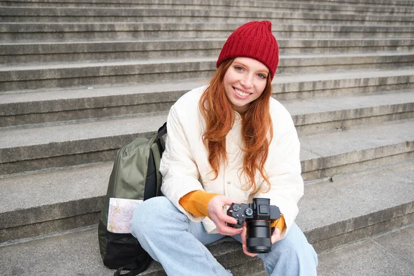 Portrait of young photographer girl, sits on stairs with professional camera, takes photos outdoors, making lifestyle shooting.