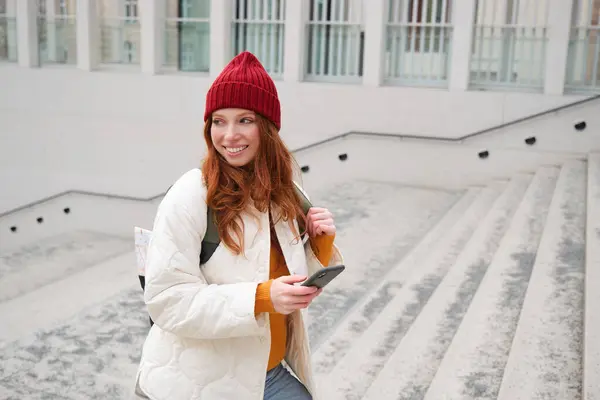Joyful girl tourist looks at mobile phone, texts message on smartphone social media application, walks around city, looks for sightseeing on mobile app.