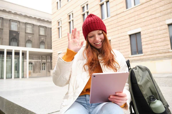 Happy redhead girl, student sits with backpack and digital tablet on street, says hello and waves at gadget camera, connects to public wifi to video chat, has online conversation.