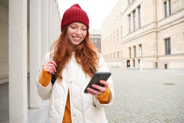Beautiful smiling girl, tourist with backpack, holding smartphone, using map on mobile phone application, looking for sightseeing in internet app, standing outdoors.