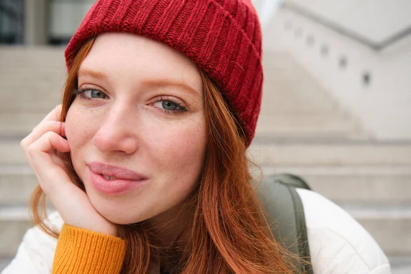 Close up portrait of beautiful redhead girl in red hat, urban woman with freckles and ginger hair, sits on stairs on street, smiles and looks gorgeous.