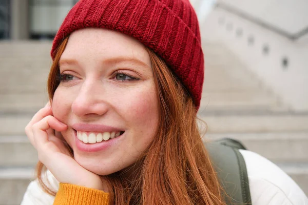 Close up portrait of beautiful redhead girl in red hat, urban woman with freckles and ginger hair, sits on stairs on street, smiles and looks gorgeous.