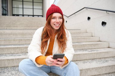 Redhead girl in red hat, sits on stairs and uses mobile phone. Modern woman holding smartphone, texting message, using telephone application outdoors.