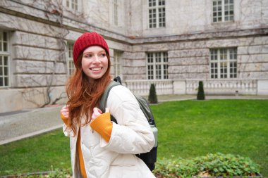 Beautiful redhead woman with backpack, tourist walking around city park and looks around, wears red hat and warm coat.