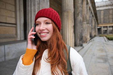 Smiling redhead female tourist talks on mobile phone and walks around city. Happy student in red hat calls friend, stands on street and uses smartphone.