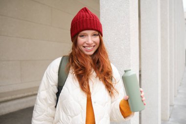 Young happy redhead woman in red hat, drinking from thermos, warming up with hot drink in her flask while walking around city, tourist relaxes with warm refreshemtn.