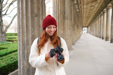 Young redhead female photographer, makes lifestyle shooting in city centre, takes photos and smiles, looks for perfect shot, makes picture.