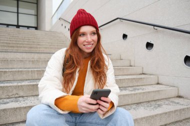 Redhead girl in red hat, sits on stairs and uses mobile phone. Modern woman holding smartphone, texting message, using telephone application outdoors.