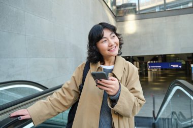 Smiling korean girl, student with smartphone goes up an escalator, reads mobile phone text message.