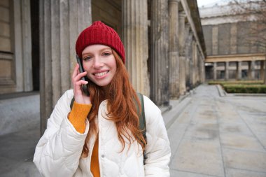Smiling redhead female tourist talks on mobile phone and walks around city. Happy student in red hat calls friend, stands on street and uses smartphone.