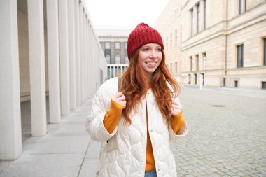 Female tourist in red hat with backpack, sightseeing, explores historical landmarks on her trip around europe, smiling and posing on street.